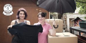 Die Geschäftsführerin von Books4Life Wien, Henriette Werner, übergibt Frau Zimmermann vom s'Häferl mehrere Kisten Sachspenden, darunter ein T-Shirt und einen Regenschirm.